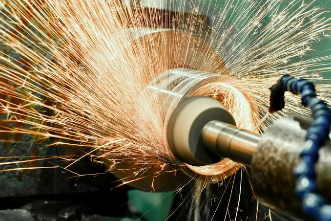 A close-up photograph of a precision CNC grinding machine processing a rotating metal cylindrical workpiece. A vibrant shower of sparks bursts forth from the machining zone, demonstrating high-speed metalwork and precision manufacturing.
